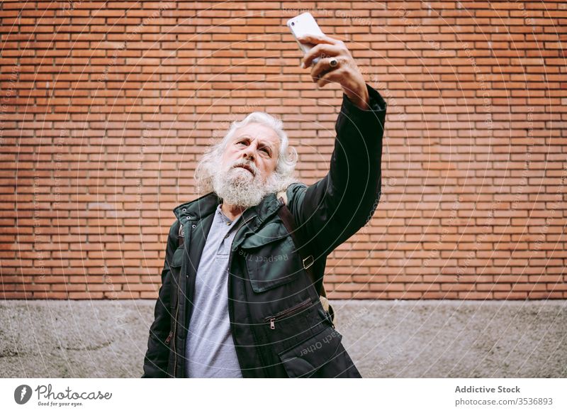 Älterer männlicher Hipster nimmt sich selbst in der Nähe einer Ziegelmauer in der Stadt Senior Selfie Mann Smartphone Großstadt ernst Backsteinwand Straße