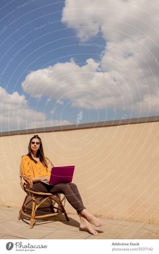 Junge Frau mit Laptop auf Stuhl sitzend Terrasse heimwärts Arbeit benutzend genießen freiberuflich Quarantäne Lifestyle Sonnenschein Browsen brünett lässig