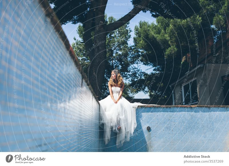 Nachdenkliche junge Braut in weißem Brautkleid ruht am Rand eines leeren Swimmingpools Frau Pool Kleid elegant Stil Garten Party Baum sinnlich träumen Sommer