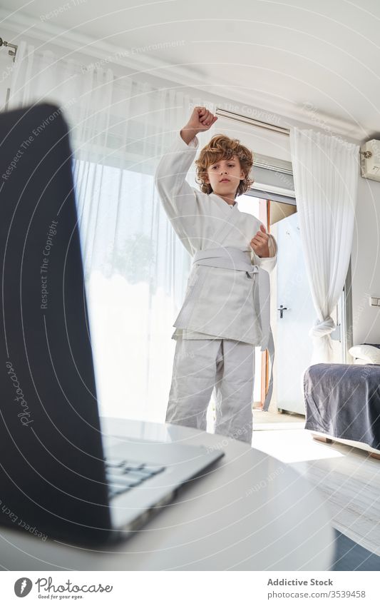 Junge in weißem Kimono lernt zu Hause am Laptop Judo jung online Tutorial heimwärts positionieren lernen üben Training Kindheit Aktivität krause Haare Computer