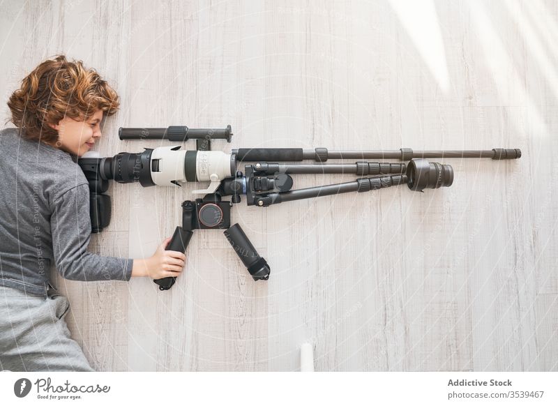 Junger Fotograf spielt mit Zubehör für professionelle Kamera wie mit Gewehr, während er auf dem Boden liegt Fotoapparat Scharfschütze Spiel so tun, als ob