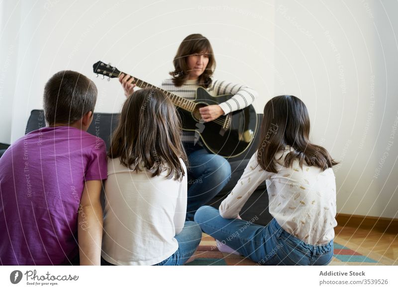 Fröhliche Frau spielt zu Hause im Wohnzimmer Gitarre für Kinder Menschengruppe Sofa heiter Raum akustisch Stock Teppich spielen Zusammensein heimwärts Freizeit