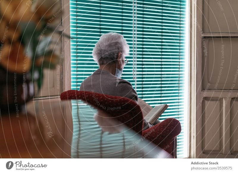 Ältere Dame mit Schutzmaske und Handschuhen liest zu Hause Buch Aufenthalt heimwärts Coronavirus Frau Senior lesen Mundschutz Fenster einsam Quarantäne