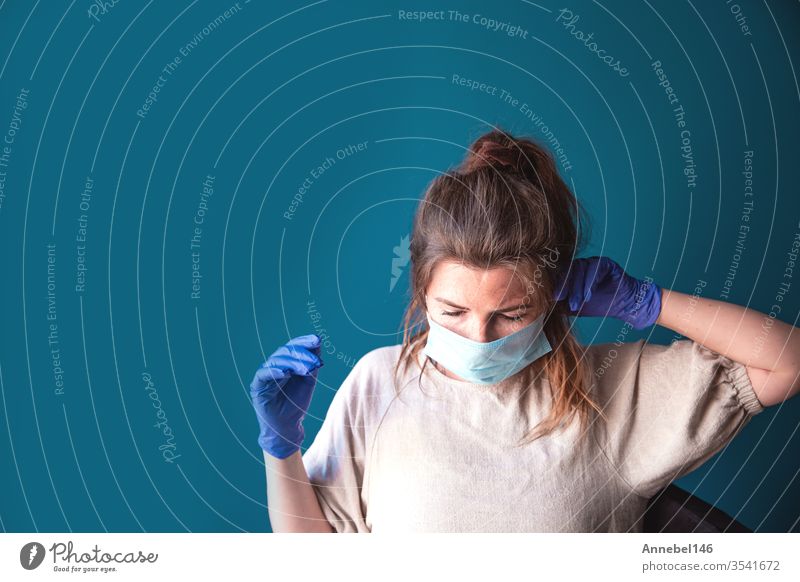 Junge Frau trägt Schutzhandschuhe und Gesichtsmaske in einem Haus in Quarantäne und sieht gelangweilt und traurig aus, für Covid-19 Coronavirus, mit blauem Hintergrund