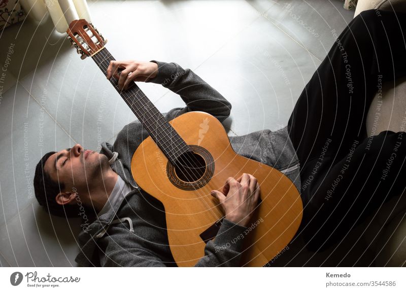 Entspannter Mann liegt auf dem Boden und spielt Gitarre in der Nähe eines hellen Fensters zu Hause. Gitarrist genießt die Musik, harmonischer Moment.