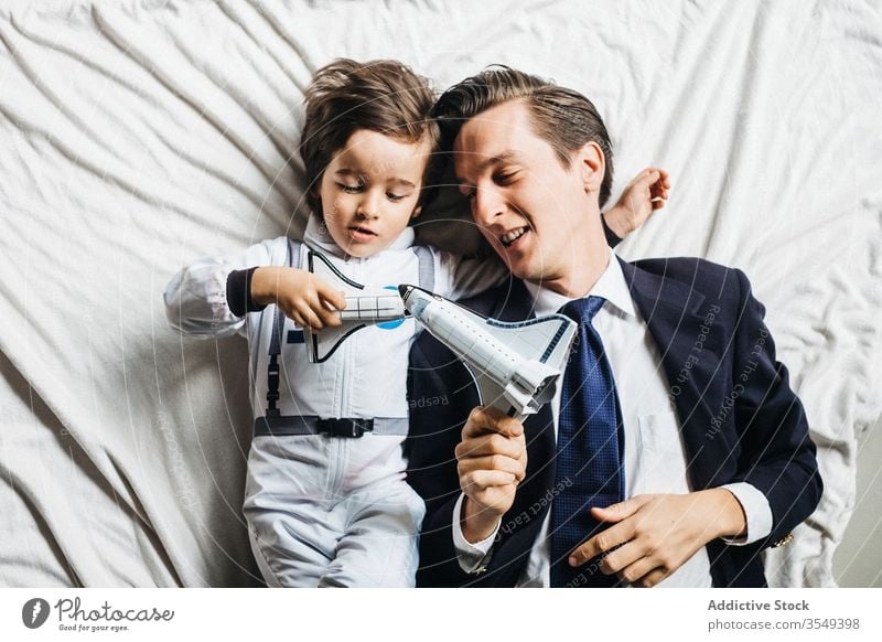 Zufriedener ethnischer Sohn und Vater liegen zusammen im Bett Astronaut Tracht Zusammensein Junge Papa spielen Spielzeug Spaß haben Lügen vielfältig