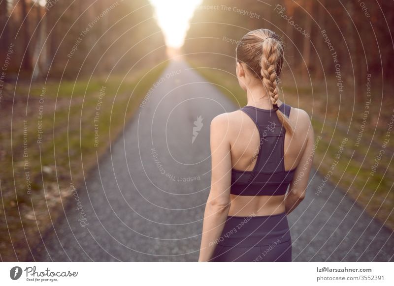 Sportlich-gesunde junge Frau, die bei Sonnenaufgang auf einer Landstraße durch einen dichten Wald dem Schein der Sonne am Ende zwischen den Bäumen in einer Nahaufnahme von hinten entgegengeht
