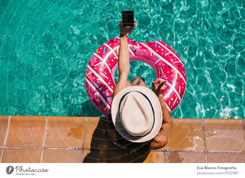 Draufsicht auf eine Frau, die sich an einem heißen sonnigen Tag im Pool mit rosa Donuts entspannt. Sommerurlaub idyllisch. Genießende sonnengebräunte Frau im Bikini und mit Hut. Ferien und sommerlicher Lebensstil. Sie benutzt ein Mobiltelefon.