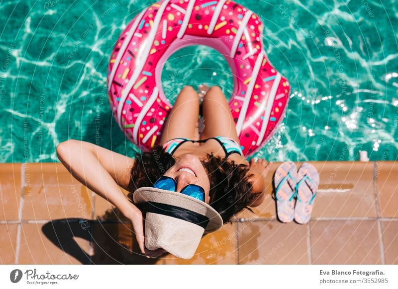 Draufsicht auf eine Frau, die sich an einem heißen sonnigen Tag im Pool mit rosa Donuts entspannt. Sommerurlaub idyllisch. Genießende sonnengebräunte Frau im Bikini und mit Hut. Urlaub und Sommer-Lifestyle