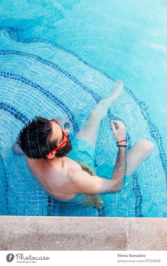 Draufsicht auf einen sexy jungen Mann mit Bart und roter Sonnenbrille, der auf den Stufen des Pools sitzt. Urlaubskonzept. Porträts Männer 1 Vollbart Spaß haben