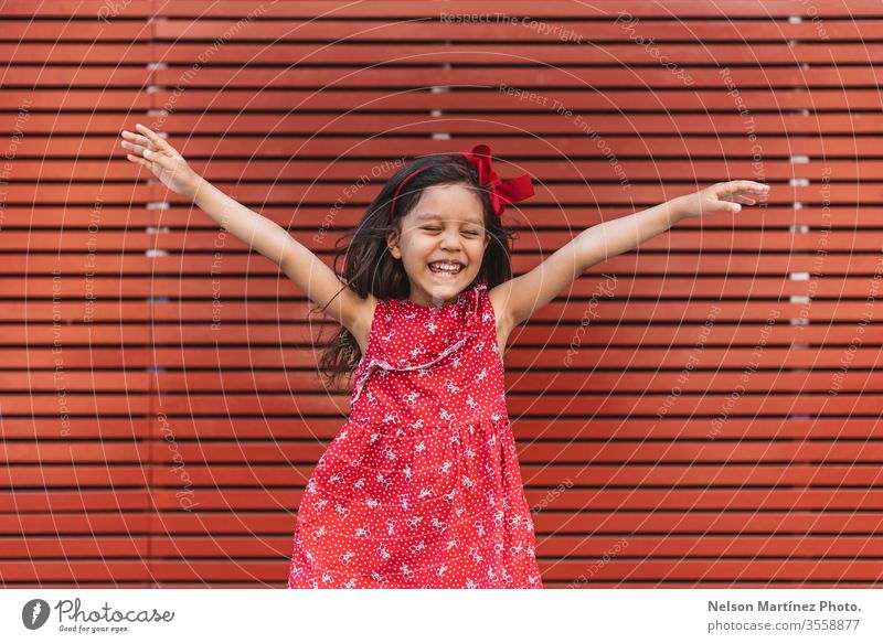 Glückliches kleines Mädchen mit weit geöffneten Armen vor rotem Hintergrund, ein rotes Kleid tragend. Sommer Spaß niedlich Kind lustig hispanisch