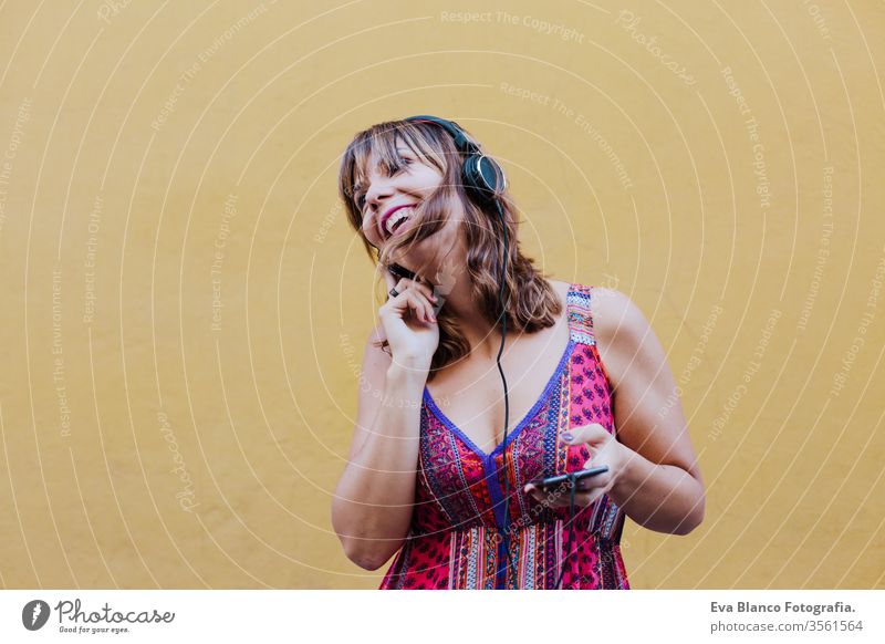 Porträt einer jungen kaukasischen Frau im Freien über gelbem Hintergrund, die über Headset und Mobiltelefon Musik hört. Sommerzeit hören Handy