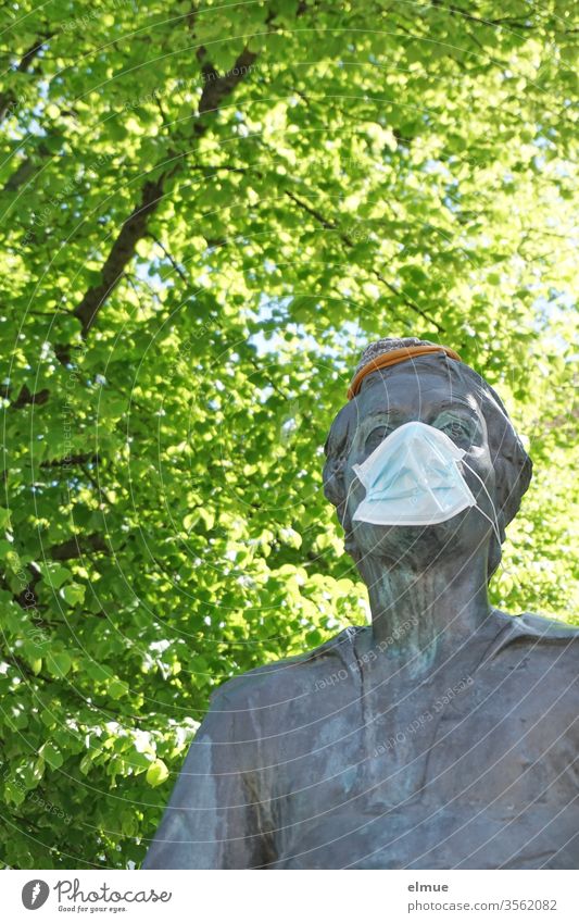 bronzene Statue von J.S.Bach mit Atemschutzmaske und Mütze unter einem grünen Baum Johann Sebastian Bach Bachdenkmal Angst Prävention Plastik verhüllen Spass