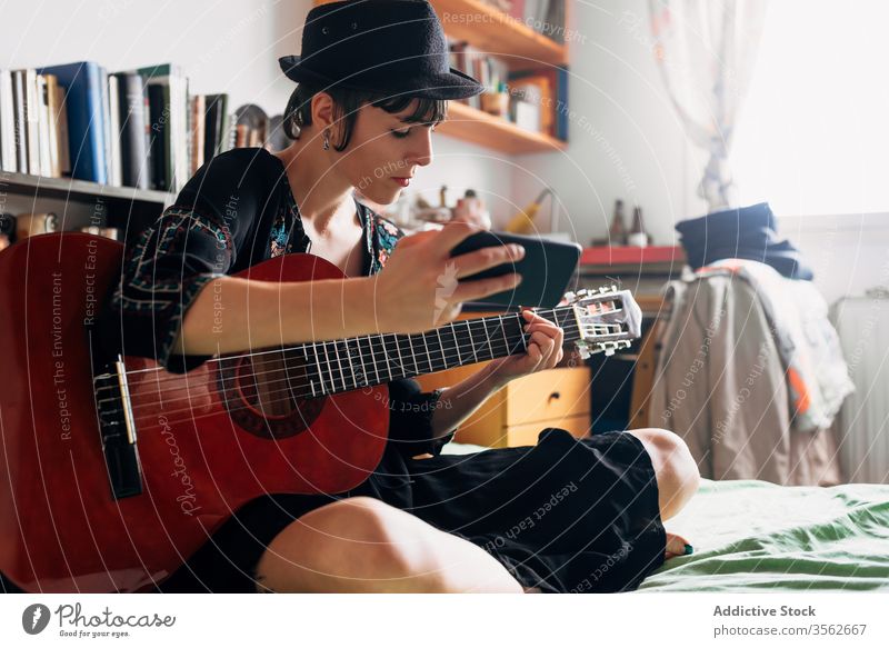 Frau mit Gitarre und Smartphone zu Hause Browsen akustisch trendy Nachricht benutzend Funktelefon Musik Wochenende Stil anhaben Hut Bett sitzen