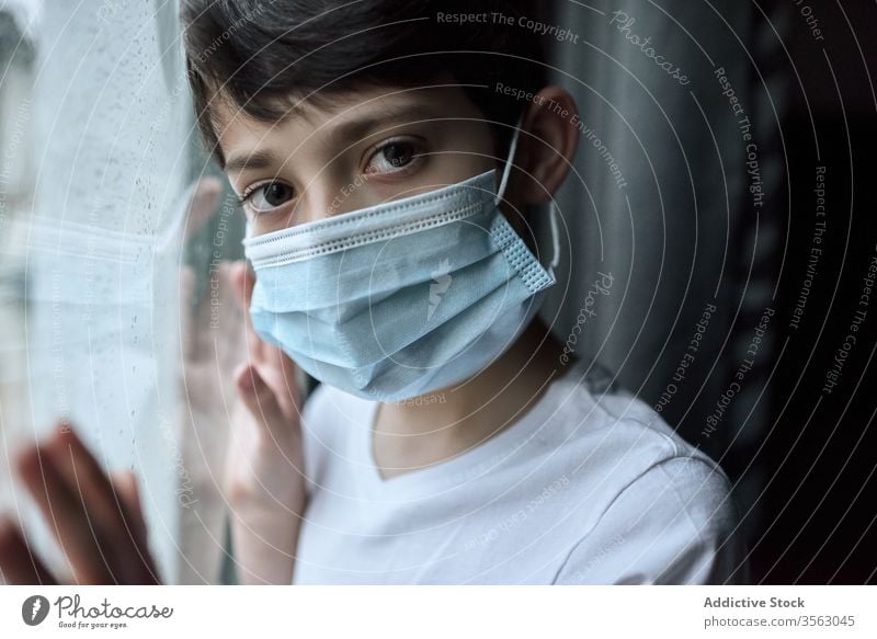 Trauriger Junge mit medizinischer Maske am Fenster Mundschutz Coronavirus Seuche heimwärts traurig unglücklich Selbstisolierung soziale Distanzierung Kind