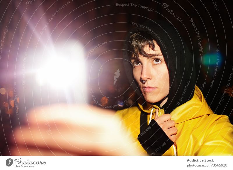 Junge seriöse Frau mit Kapuze fotografiert mit Smartphone in dunkler Nacht Licht Rochen Fotografie verwenden ernst Regenmantel schießen ruhig Vorschein reisen