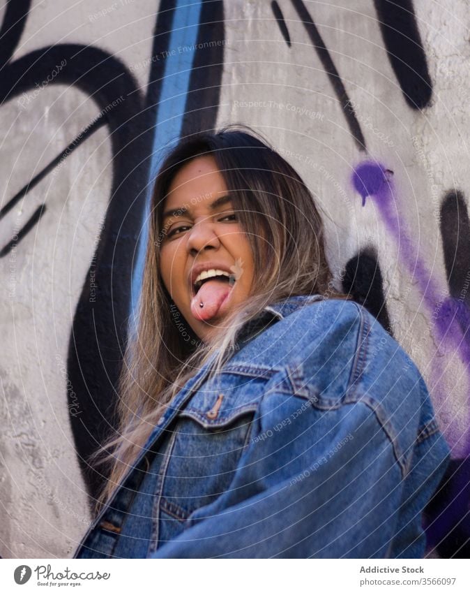 Freche ethnische Tausendjährige mit Zunge auf der Straße ausspannen tausendjährig Frau auflehnen unanständig Piercing Graffiti Wand Hipster Jeansstoff Jacke