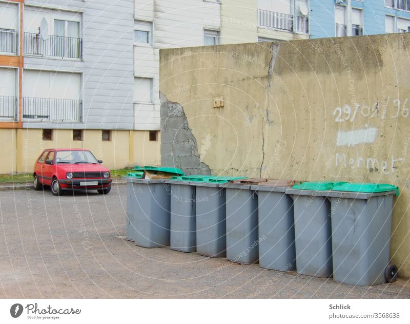 Alter Wohnblock mit rotem Auto und einer Reihe Mülltonnen mit grünen Deckeln alt Mauer Datum Mehmet Text marode trist gedeckte Farben PKW Asphalt ohne Himmel