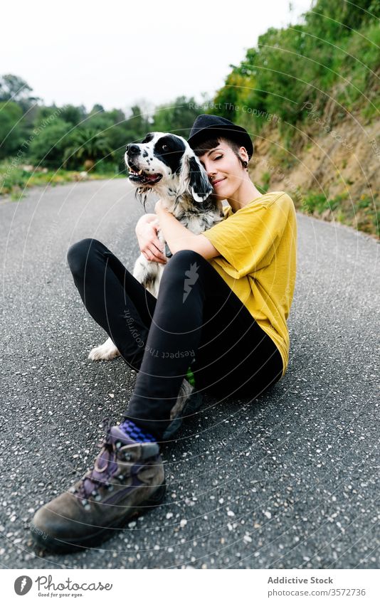 Fröhliche junge Frau sitzt mit ihrem Hund am Boden heiter Spaziergang Spaß Park Natur Hipster Subkultur Stil Zusammensein Begleiter Straße Hut Haustier Freizeit