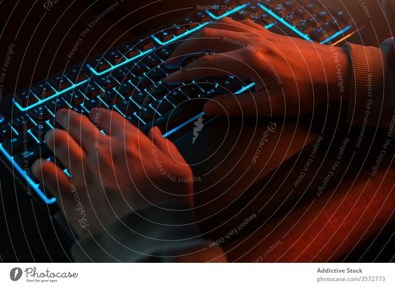 Crop-Programmierer, der zu Hause in einem dunklen Raum mit blauer Hintergrundbeleuchtung auf einer Tastatur tippt Keyboard Tippen Nacht Hand Mann Komfort dunkel