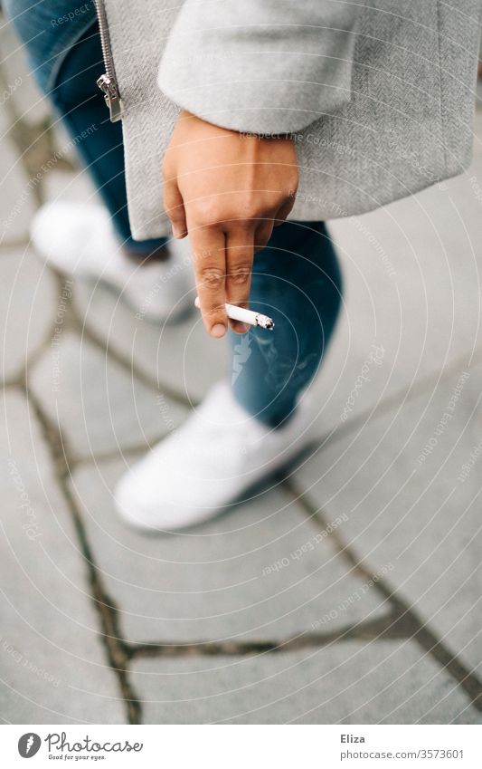 Frau beim Rauchen mit Zigarette in der Hand Nikotin Sneaker Jeans draußen brennend Tabakwaren ungesund Sucht Nikotingeruch Suchtverhalten Abhängigkeit hell weiß