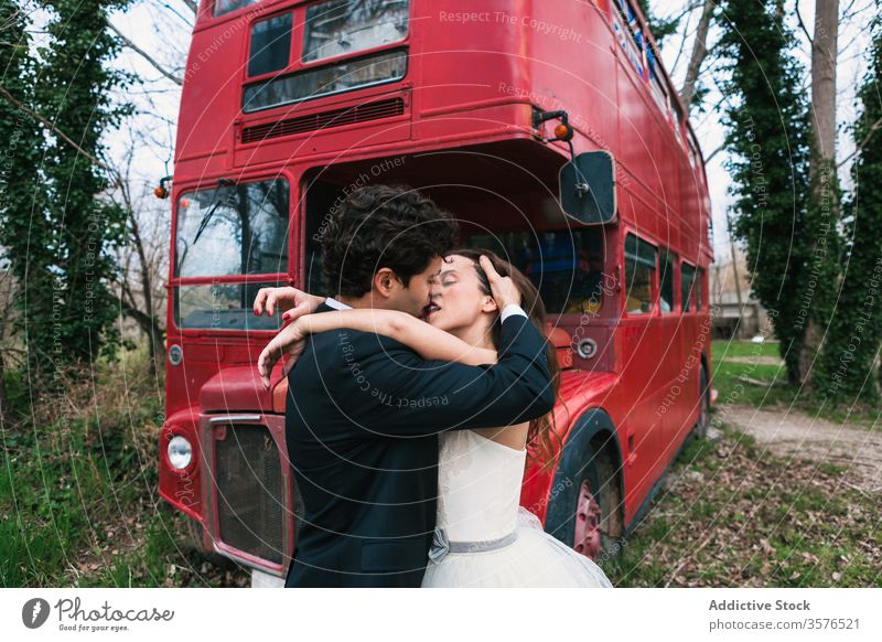 Stilvolles frisch verheiratetes Paar umarmt und küsst sich in der Nähe eines Doppeldeckerbusses Jungvermählter Kuss Umarmung roter Bus Wald Hochzeit Umarmen