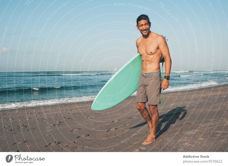 Fröhlicher Surfer mit Surfbrett am Strand stehend Mann MEER Glück aktiv Meer Küste Lächeln ohne Hemd ethnisch männlich Lifestyle Sand Sommer Ufer Urlaub Wasser