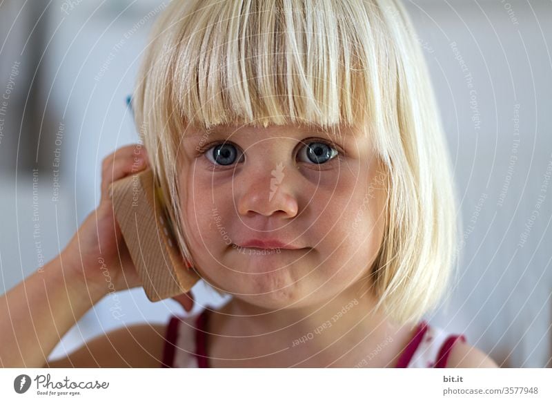 Niedliches, süßes, blondes Mädchen hält Spielzeug-Telefon in der Hand und wartet neugierung, gespannt auf Freizeichen. Natürliches, fröhliches Kind spielt mit Holztelefon telefonieren. Biologisches, ökologisches, nachhaltiges, ungiftiges Kinderspielzeug.