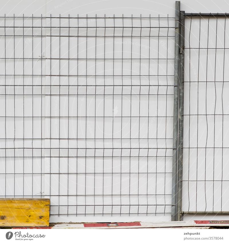 Ein Bauzaun for weisser Wand. am Boden liegen Verschalungsbretter und Absperrbretter rechtwinklig Rechtecke Gitter Strukturen & Formen Bretter weiß grau gelb