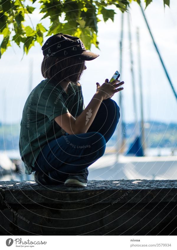 Junge spielt auf dem Handy am Hafen young praktisch Smartphone sitzen Sommer Spiel spielen Verschlussdeckel Schatten blau Natur pflanze sehen Landschaft