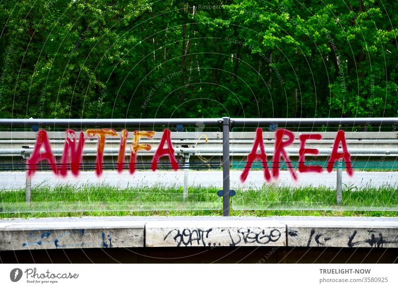 Wörtlich genommen: | Frohe Botschaft...  Graffiti ANTIFA AREA in roten Grossbuchstaben auf durchsichtiger Trennwand einer Tram Haltestelle an Schnellstraße mit Wald im Hintergrund