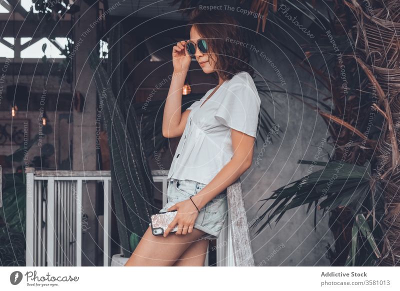Selbstbewusste junge Frau mit Sonnenbrille auf der Terrasse gegen exotische Pflanzen im Hotel selbstbewusst Lächeln Tourismus Loft Smartphone cool tropisch Bali