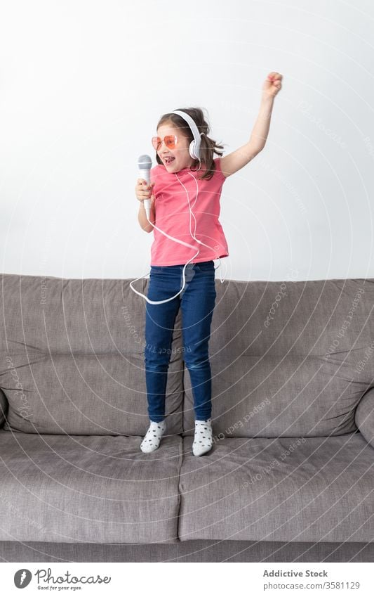 Glückliches Mädchen im Vorschulalter mit Kopfhörern und Mikrofon singt, während sie alleine zu Hause spielt singen Spaß haben Kind springen Sofa heimwärts