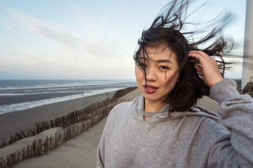 Junge Asiatin steht bei windigem Wetter am Meeresstrand Frau MEER Strand Skyline Harmonie Sand Haare berühren Küste nachdenklich reflektierend Windstille