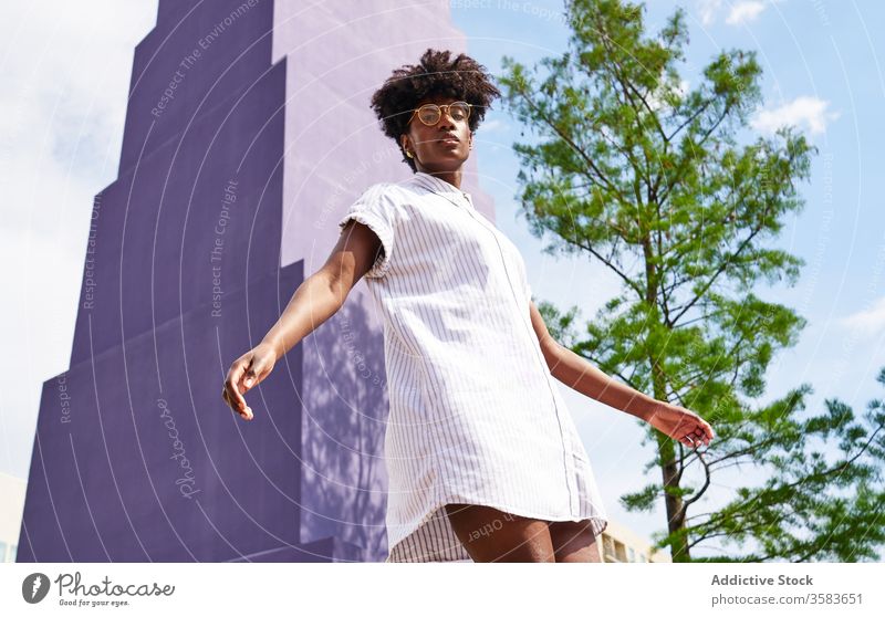 Ruhige schwarze Frau auf der Straße vor bunt violetter Fassade eines Wolkenkratzers und grünem Baum in der Stadt Kleid Vorschein Persönlichkeit Windstille