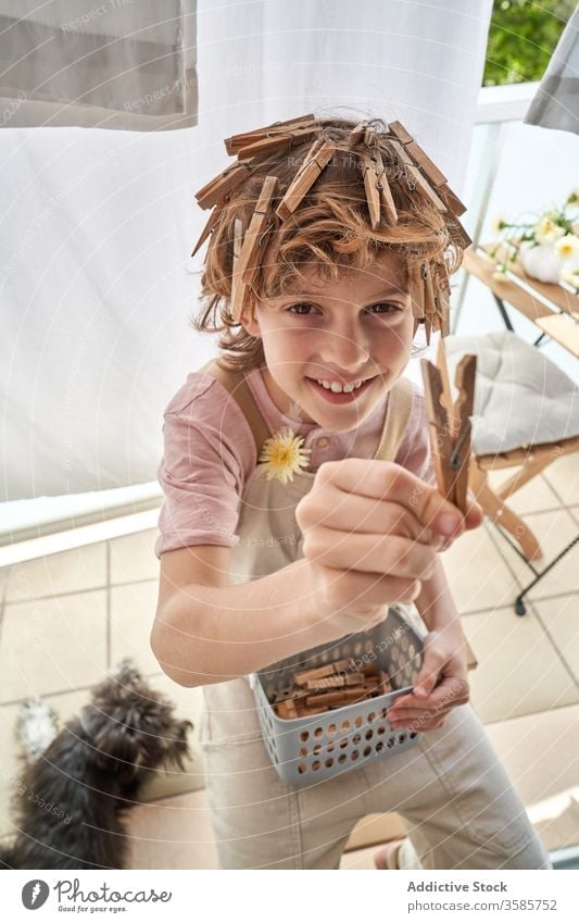 Fröhlicher kleiner Junge mit an den Haaren befestigten Wäscheklammern auf dem Balkon sitzend heimwärts Spaß Kleiderspin Frisur so tun, als ob Spiel Glück