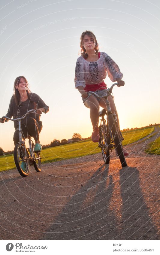 Zwei modische, hübsche Teenager fahren gemeinsam auf alten Rädern in der Abendsonne um die Wette. 2 feminine Jugendliche auf Fahrradtour draussen in der Natur auf Straße. Schwestern auf Wettfahrt mit nostalgischen Fahrrädern vor grüner Wiese im Urlaub.