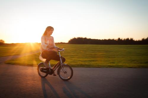 Langhaarige, brünette Jugendliche, fährt bei Abendlicht mit einem kleinen Fahrrad auf der Straße. Fahrradtour in der Dämmerung, vorbei an grüner Wiese und Wald, wobei der Teenager, wie ein lustiger Riese auf dem überdimensional, winzigen Zwergen-Rad wirkt.