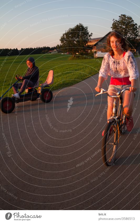 Mutter und hübsche Tochter fahren gemeinsam auf altem Rad und Kettcar in der Abendsonne um die Wette. Jugendliche und Erwachsene auf Fahrradtour draussen in der Natur. Lustige Wettfahrt mit nostalgischen Fahrzeugen vor grüner Wiese im Urlaub auf Bauernhof.