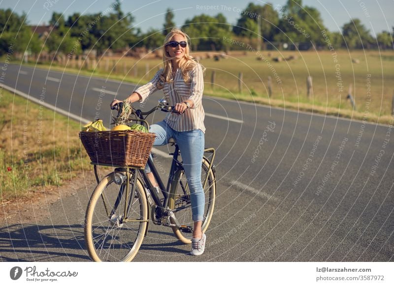 Eine blonde Frau mittleren Alters, die auf ihrem Fahrrad Lebensmittel einkaufte, hielt am Straßenrand mit einem Korb voller gesunder frischer Produkte an, während sie lächelnd in die Frühlingssonne in einer ländlichen Landschaft blickte