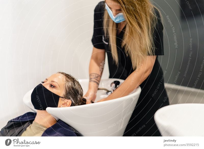 Friseurin, die in einem Salon den Kopf eines Kunden wäscht, geschützt durch eine Maske Waschen Behaarung Frau Massage Waschbecken Stylist Schönheit