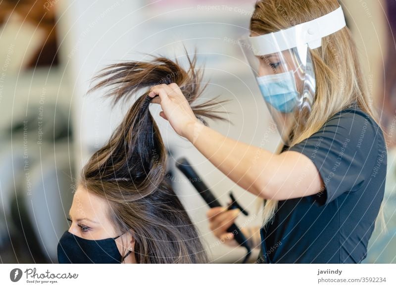 Friseurin, durch eine Maske geschützt, kämmt in einem Salon die Haare ihrer Kundin mit einem Bügeleisen. Frau bügeln Wellen Behaarung Mädchen Frisur Pflege