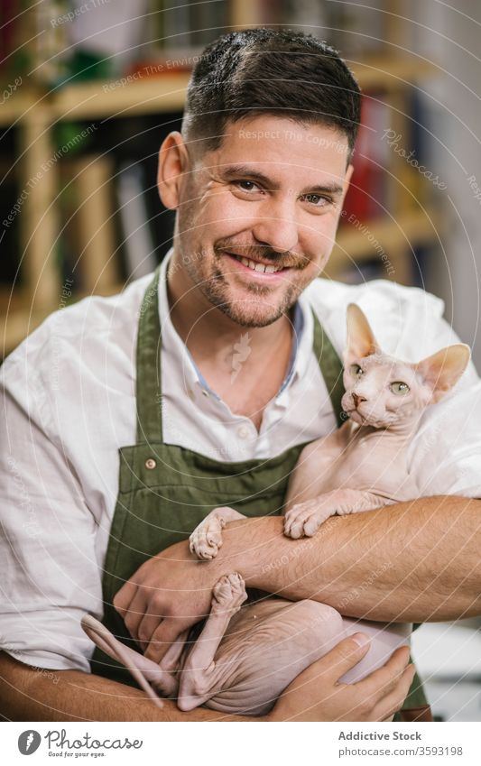 Glücklicher erwachsener Mann mit Sphynx-Katze im Workshop Werkstatt Kunstgewerbler Lächeln Arbeitsraum Haustier Uniform Atelier haarlos Begleiter Handwerker