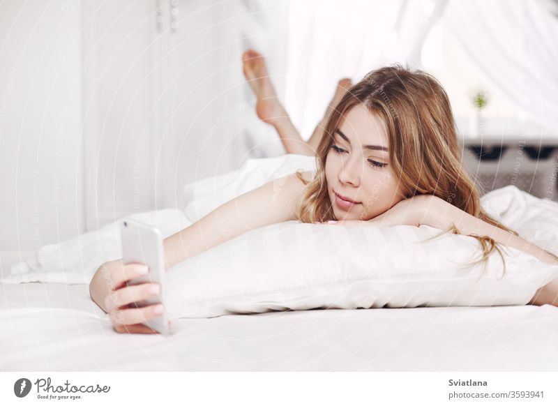 Ein süßes, lächelndes Mädchen schaut auf das Telefon, das auf dem Bett liegt. Ein Mädchen liest Nachrichten auf ihrem Telefon, wenn sie morgens aufwacht. Frau