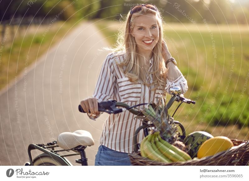 Frau, die ihr Fahrrad benutzt, um frische Produkte zu kaufen, steht auf einer Landstraße, die von einem warmen Sonnenstrahl beleuchtet wird, der fröhlich in die Kamera lächelt, während sie ihr Fahrrad mit einem Korb voller Obst und Gemüse hält