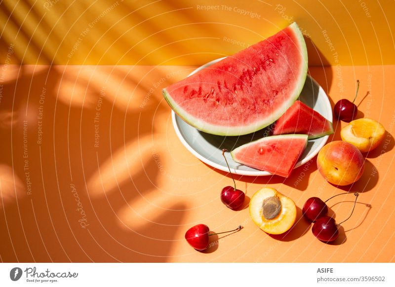 Sommerfruchtkomposition mit Schatten tropischer Blätter auf orangem Hintergrund Frucht farbenfroh Wassermelone Stillleben Handfläche Kirsche Aprikose gelb