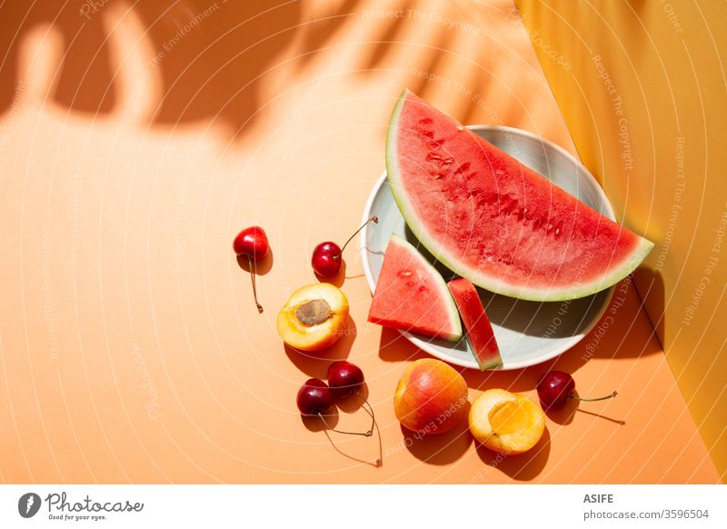 Sommerfruchtkomposition mit Schatten tropischer Blätter auf orangem Hintergrund Frucht farbenfroh Wassermelone Stillleben Handfläche Kirsche Aprikose gelb