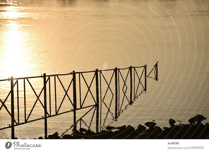 Ein Geländer führt in einem Fluss in dem sich das abendliche Sonnenlicht spiegelt Metall Linie Gegenlicht Konstruktion Wasser Flussufer Sonnenlichtstrahlen