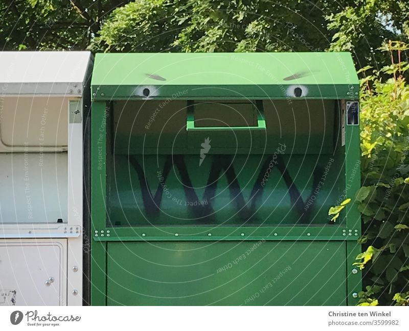 Bitte recht freundlich / Grüner Altkleider Container mit aufgemaltem Gesicht steht im Sonnenschein vor grünen Sträuchern und Bäumen Altkleidercontainer