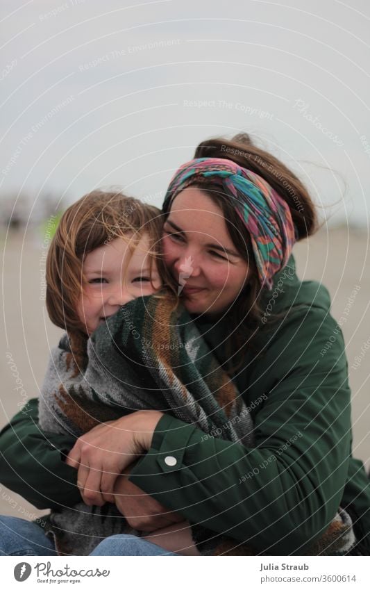 Mutter und Kind am Strand bei frischer Brise Regenjacke Haarband Decke Ethnomuster grün Wind festhalten drücken Jeanshose lachend freuen genießen Kleinkind
